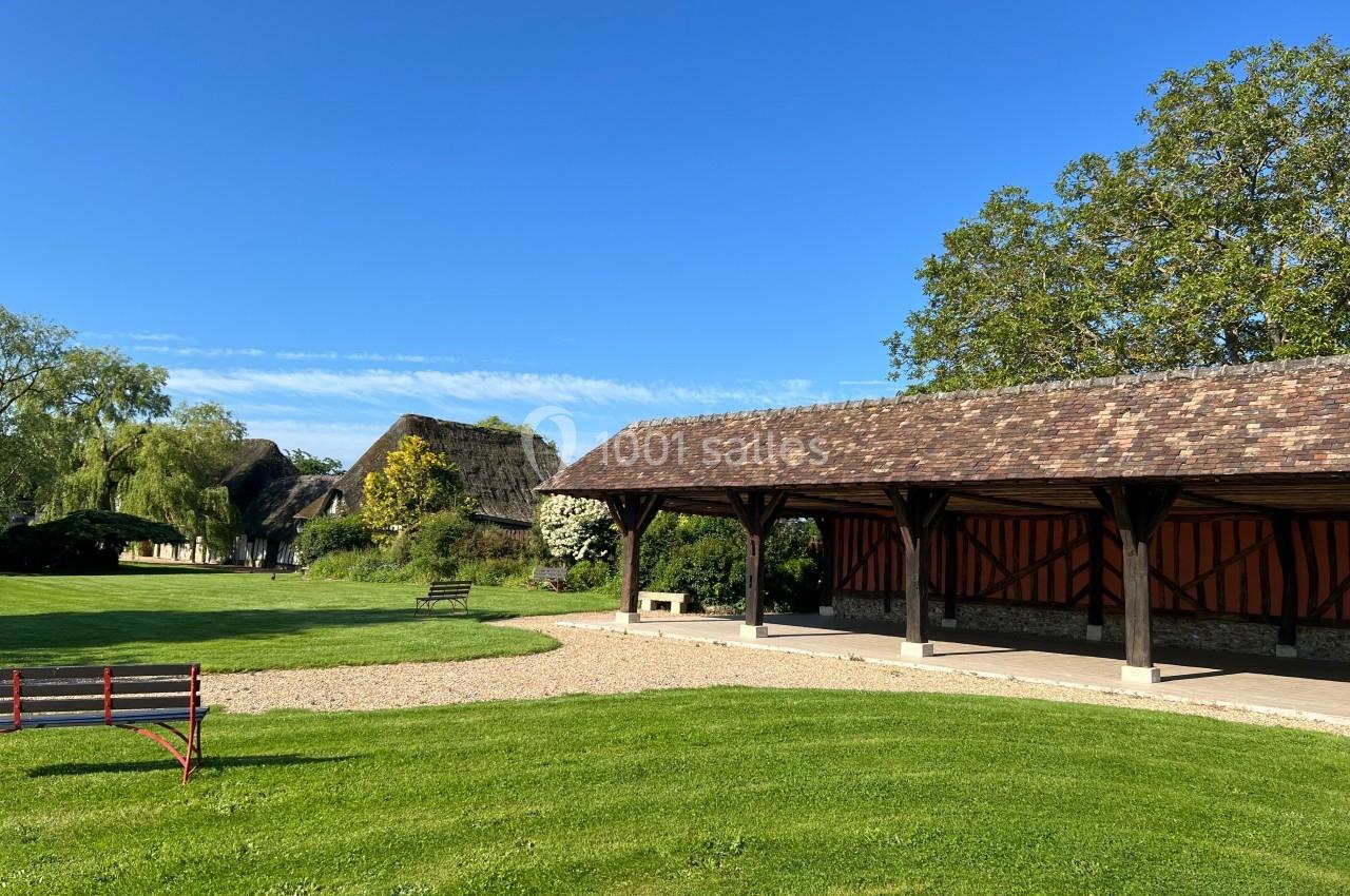 Abri en bois ouvert sur un espace vert avec des bâtiments à toit de chaume en arrière-plan sous un ciel bleu.