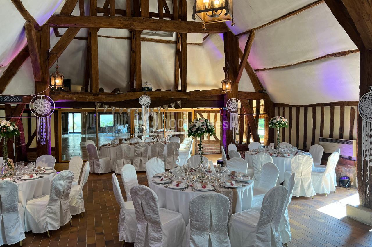 Salle de réception rustique avec poutres apparentes, tables rondes dressées et décorations élégantes.