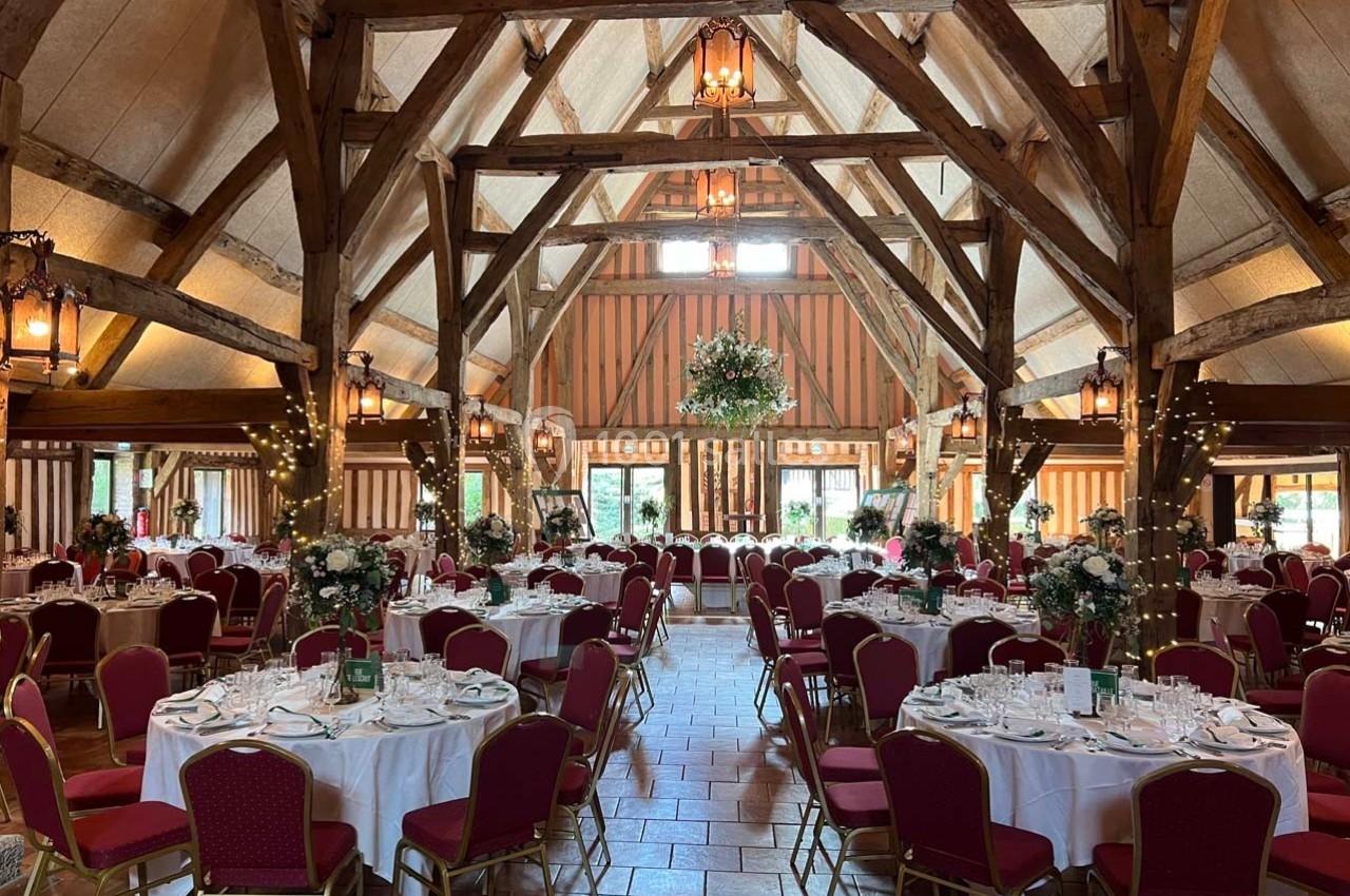 Salle de réception avec poutres apparentes, tables dressées avec nappes blanches, chaises rouges et décor floral.