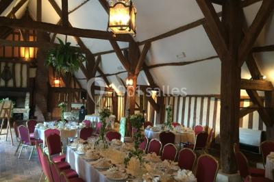Salle de réception rustique avec poutres en bois apparentes, tables rondes dressées et chaises rouges.