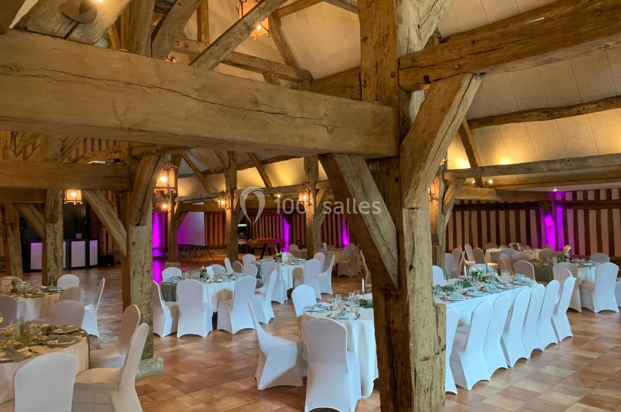 Salle de réception rustique avec poutres apparentes, tables rondes dressées et chaises blanches, éclairage tamisé.