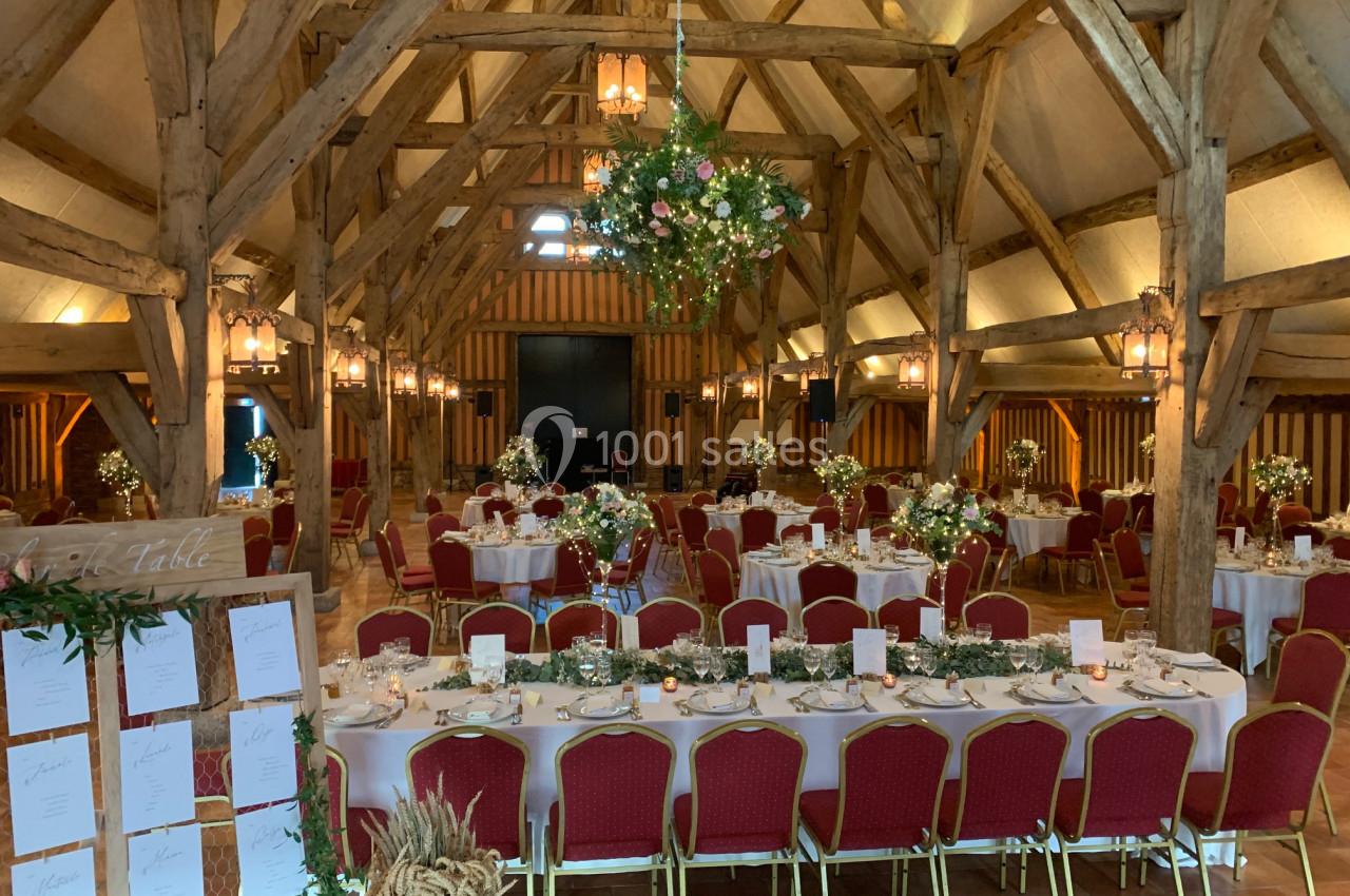 Salle de réception rustique avec charpente en bois, tables décorées de fleurs et chaises rouges alignées.