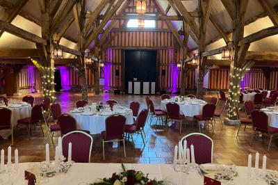 Salle de réception rustique avec poutres en bois apparentes, tables rondes dressées et chaises rouges.