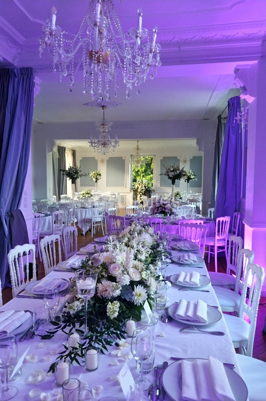 Salle de réception élégante décorée pour un mariage, avec tables dressées, fleurs blanches et éclairage violet.