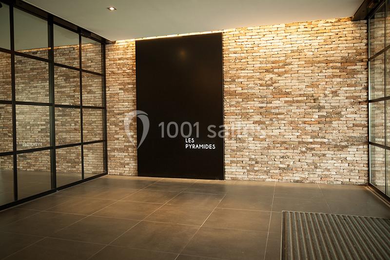Mur en briques apparentes avec une plaque noire portant l'inscription ’Les Pyramides’, encadré par des parois vitrées.