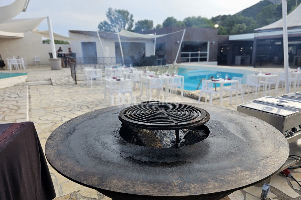 Plancha ronde en métal avec grille au premier plan, située près d'une piscine entourée de tables et chaises blanches.
