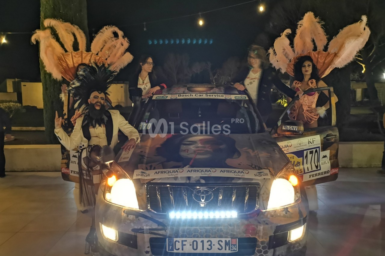 Quatre personnes en costumes extravagants posent devant un véhicule décoré, éclairé par des phares allumés, de nuit.