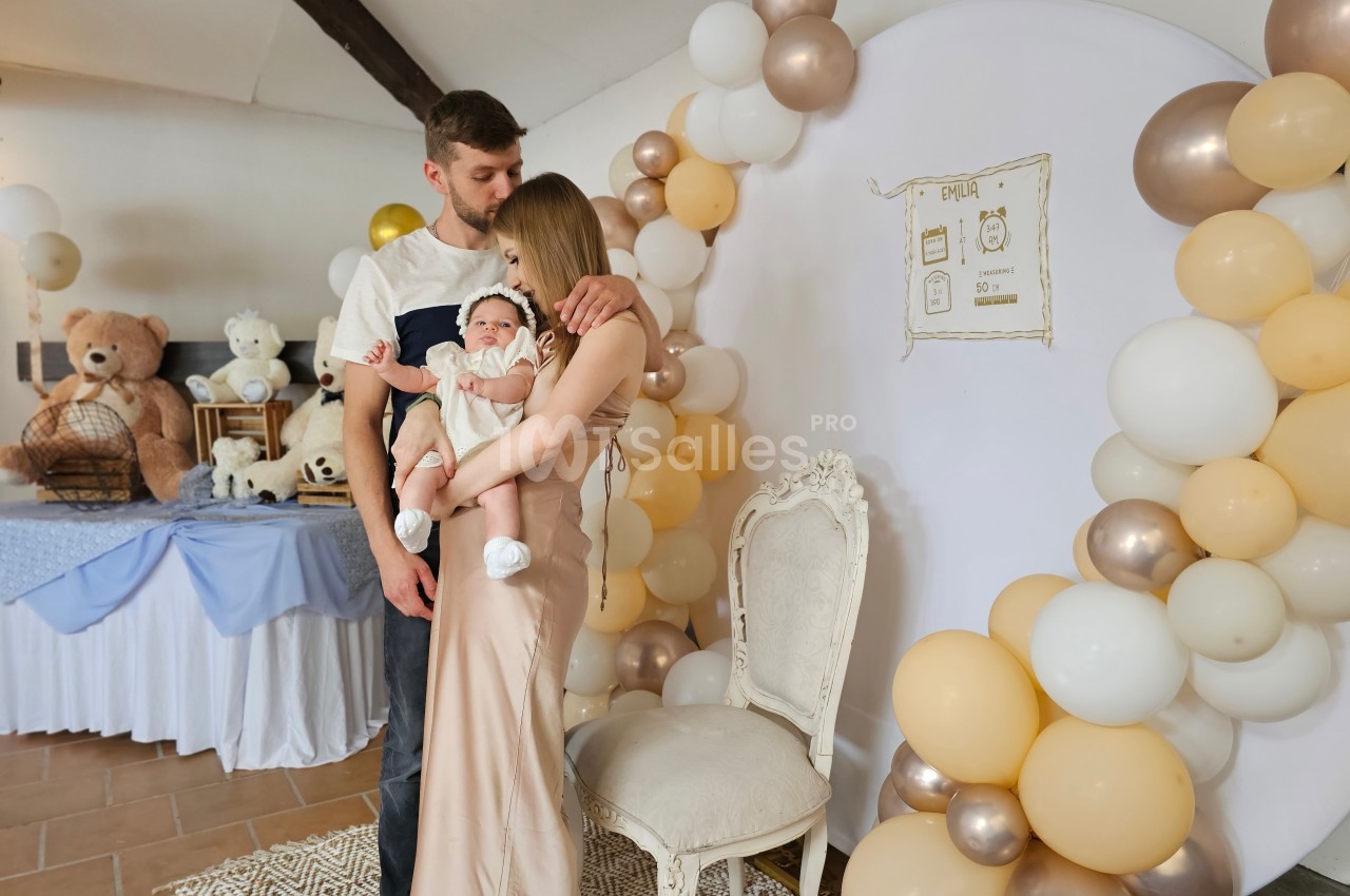 Un couple tenant un bébé dans une salle décorée avec des ballons et des éléments sur le thème de l'enfance.