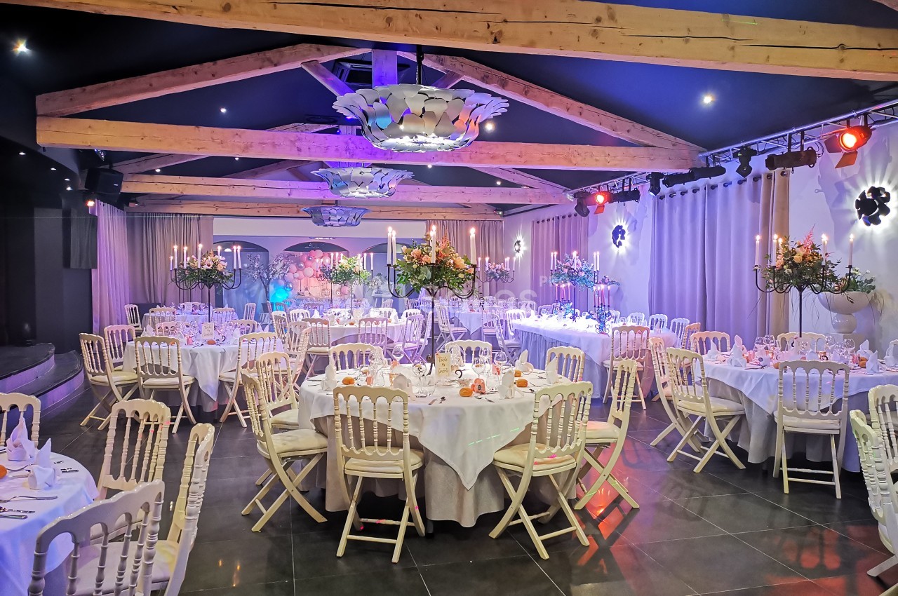 Salle de réception élégante avec tables rondes dressées, chaises blanches et décorations florales sous des poutres…
