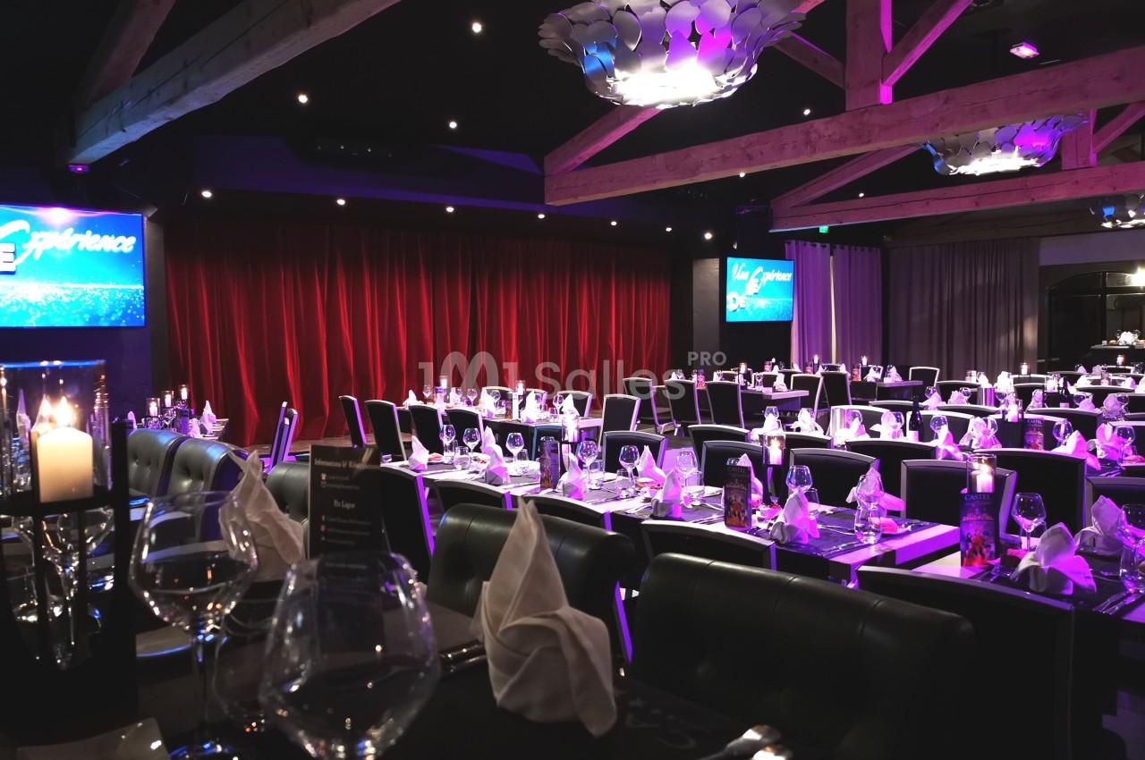 Salle de spectacle avec tables dressées, nappes blanches, chandeliers et éclairage tamisé, rideaux rouges en fond.