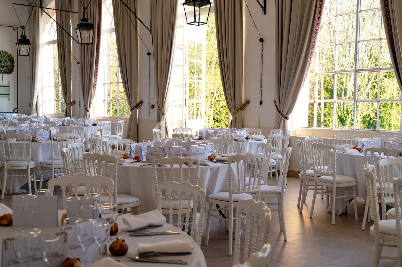 Salle de réception lumineuse avec tables rondes dressées, nappes blanches et chaises en bois clair, près de grandes fenêtres.
