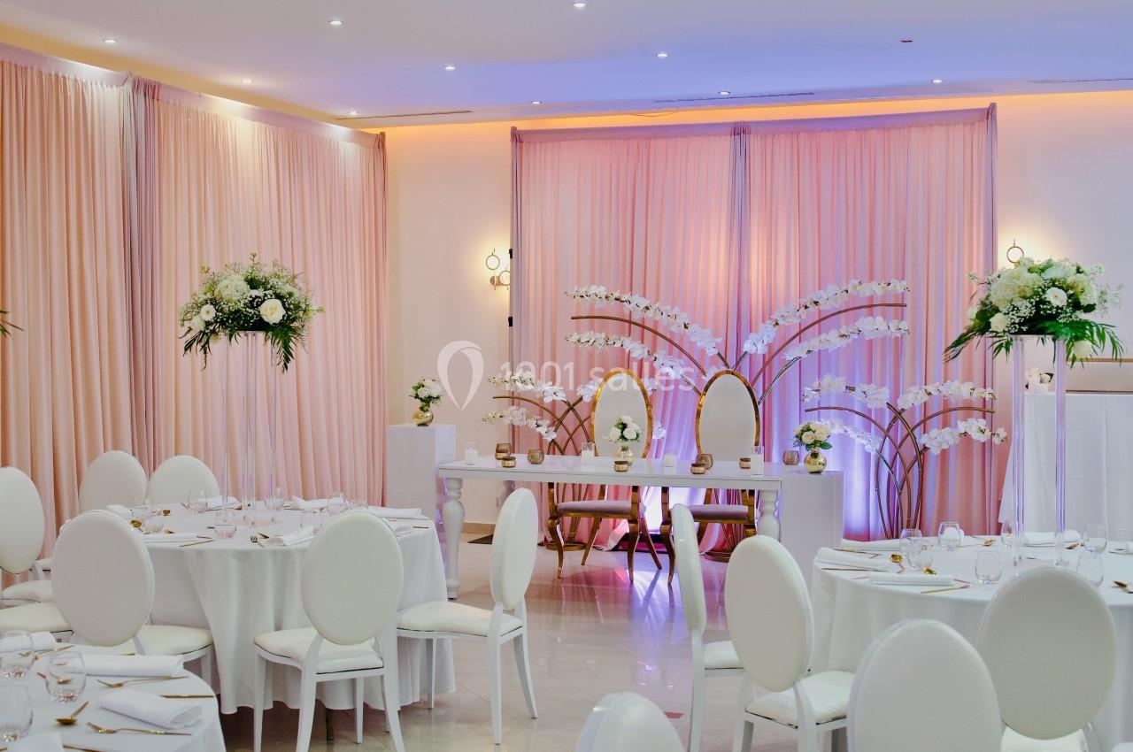 Salle de réception décorée avec des tables rondes, des chaises blanches et des arrangements floraux élégants.