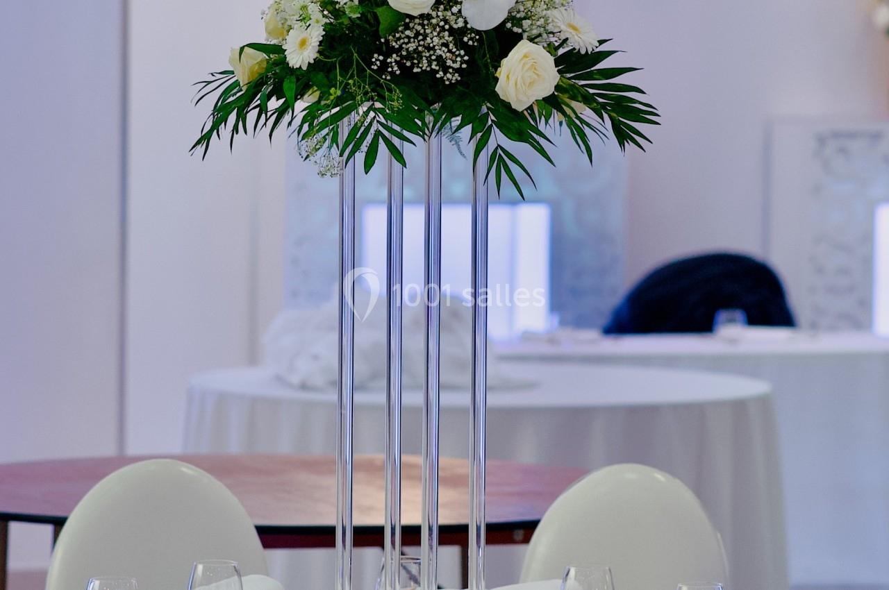 Centre de table floral avec roses blanches et feuillage, posé sur un support haut dans une salle de réception élégante.