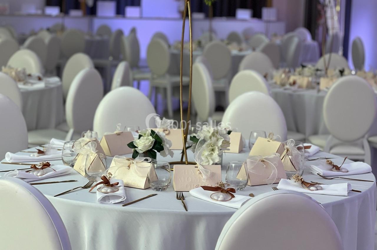 Table ronde élégamment dressée avec nappes blanches, décorations florales et cadeaux d'invités dans une salle de réception.