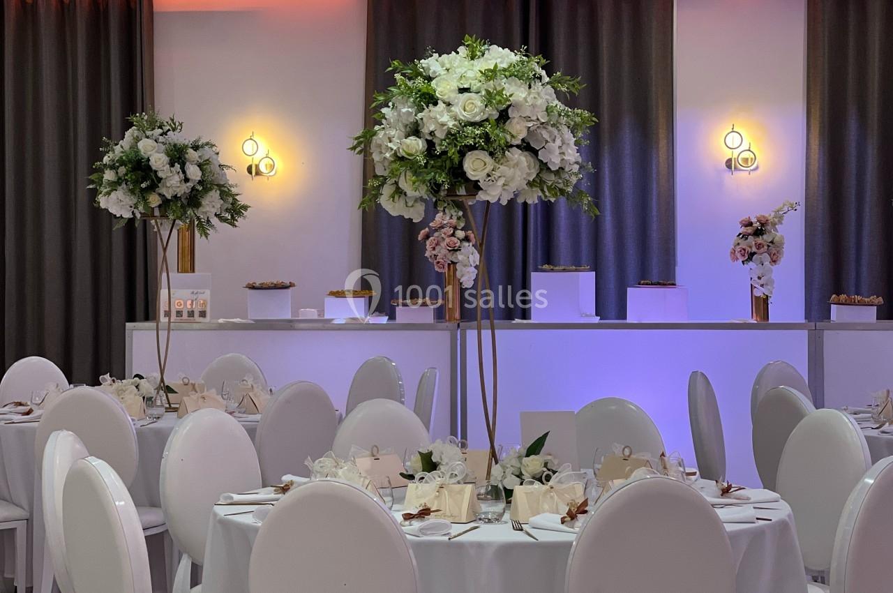 Salle de réception décorée avec des tables rondes, des chaises blanches et des centres de table fleuris.