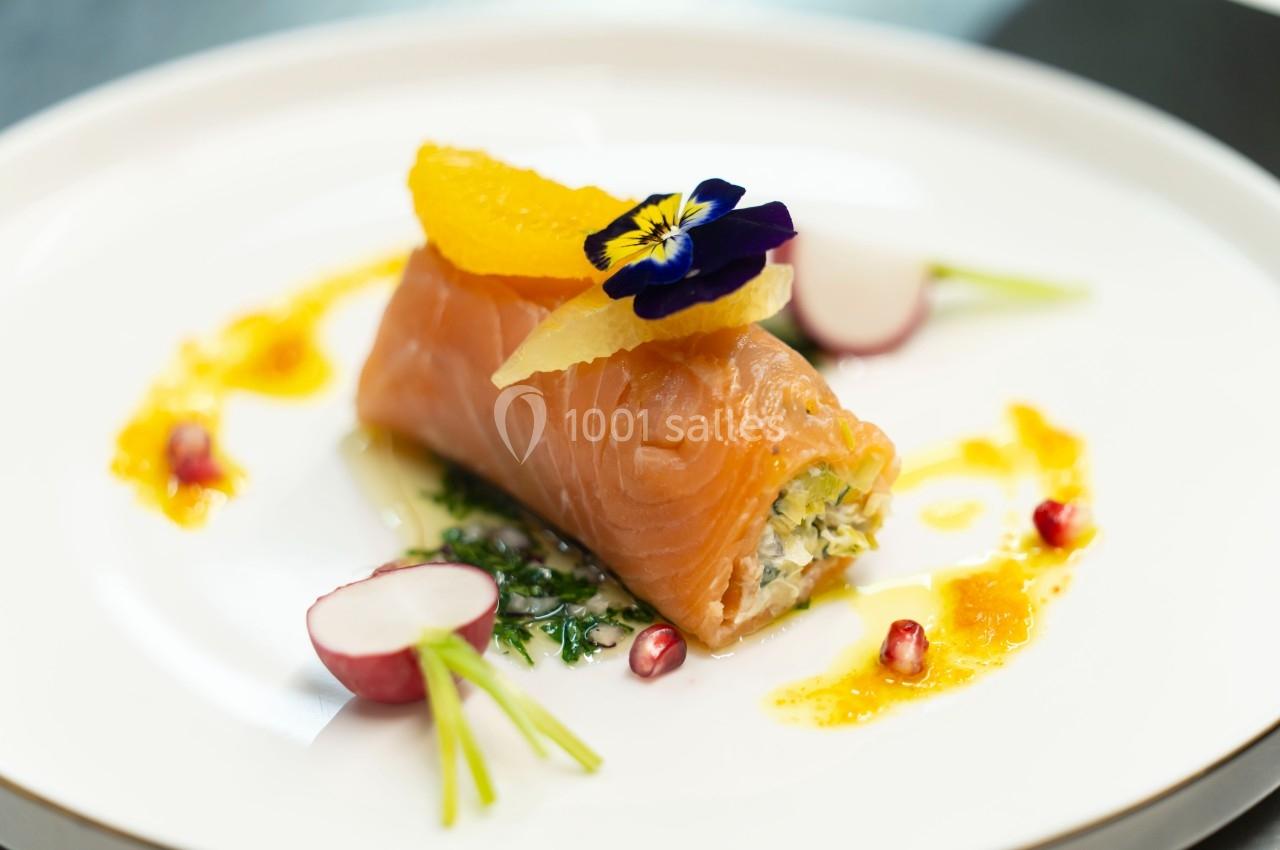 Rouleau de saumon fumé garni, décoré de fleurs comestibles, tranches d'orange et radis sur une assiette blanche.