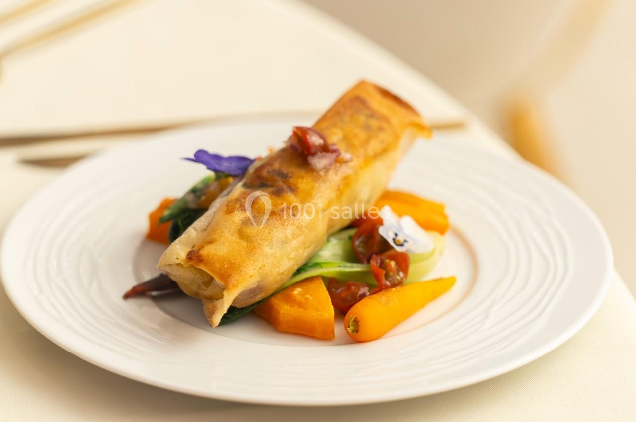 Rouleau croustillant servi sur un lit de légumes colorés avec une garniture florale sur une assiette blanche.