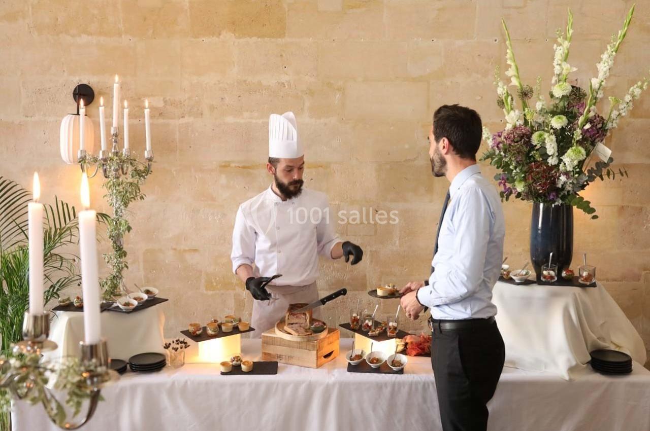 Location salle Chantilly (Oise) - La Capitainerie du Château de Chantilly #3 Un chef en tenue blanche prépare des plats devant un invité à une table décorée de fleurs et de chandeliers.