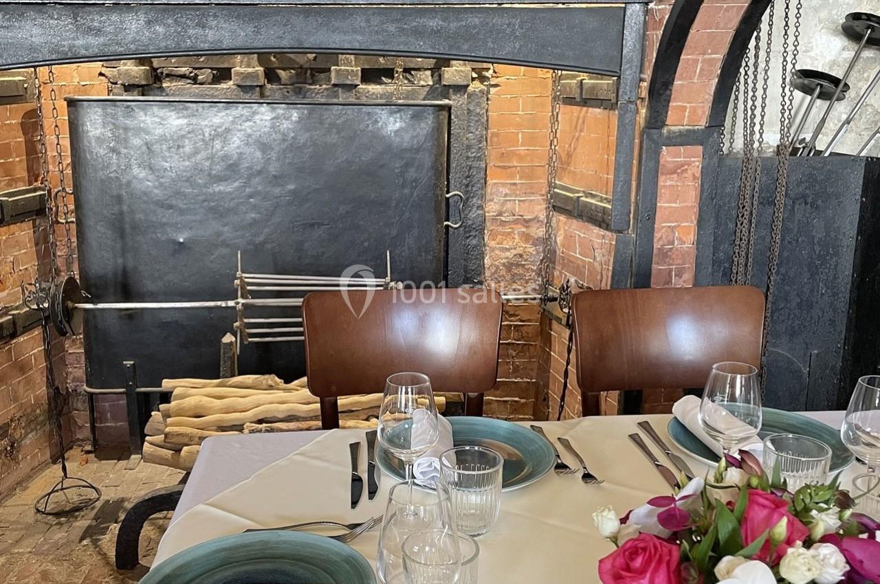 Location salle Chantilly (Oise) - La Capitainerie du Château de Chantilly #17 Table dressée avec vaisselle et fleurs devant une cheminée en briques avec des outils de foyer suspendus.