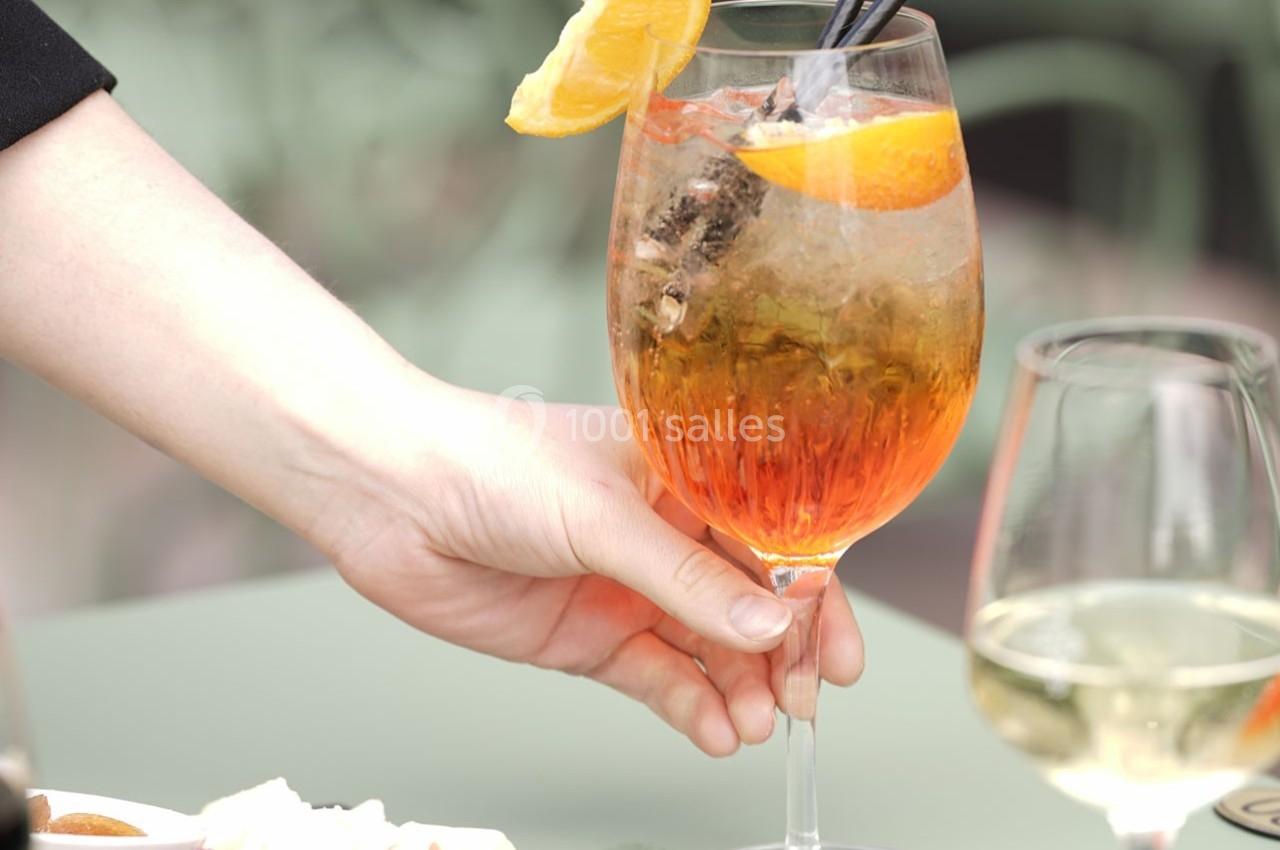 Location salle Chantilly (Oise) - La Capitainerie du Château de Chantilly #21 Un verre de cocktail orange garni d'une tranche d'orange, tenu par une main sur une table verte.