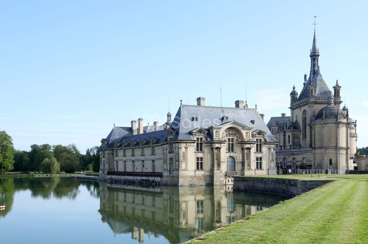 Façade d'un château historique bordé par un plan d'eau, entouré de pelouses et d'arbres sous un ciel dégagé.
