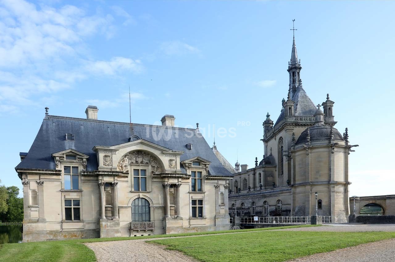 Façade d'un château historique en pierre avec des toits d'ardoise, entouré de pelouse et sous un ciel dégagé.