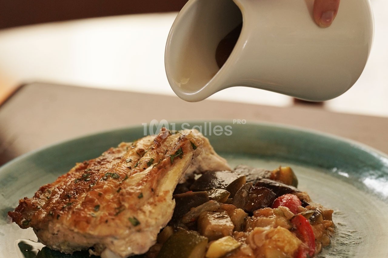Une main verse une sauce brune depuis un petit pichet sur un plat de poulet grillé accompagné de légumes rôtis.