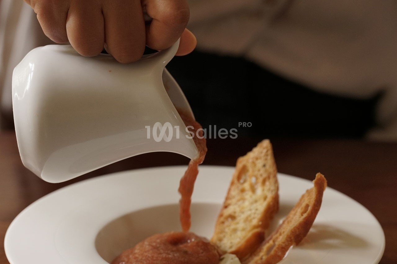 Une main verse une sauce rouge depuis un petit pichet blanc dans une assiette contenant des croûtons.