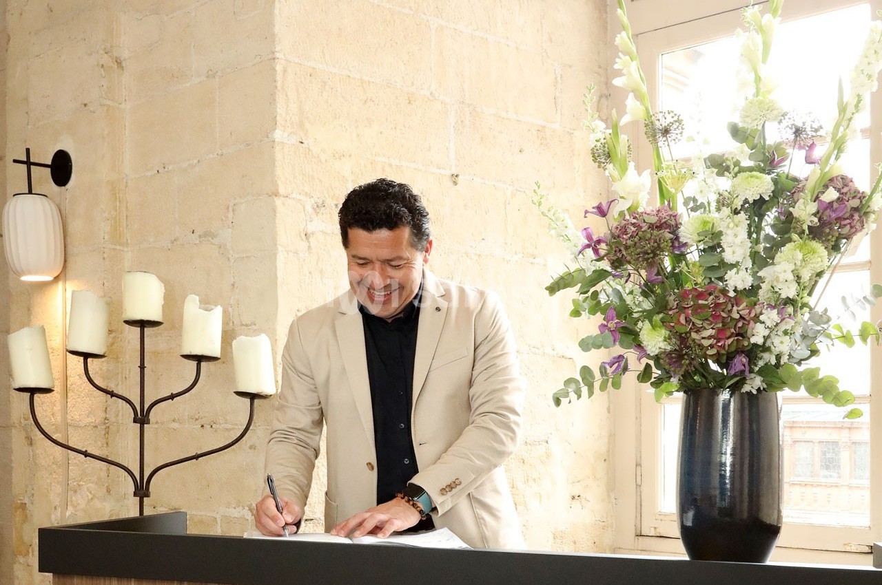 Un homme souriant signe un document à un comptoir, près d'un vase de fleurs et d'un chandelier à bougies.