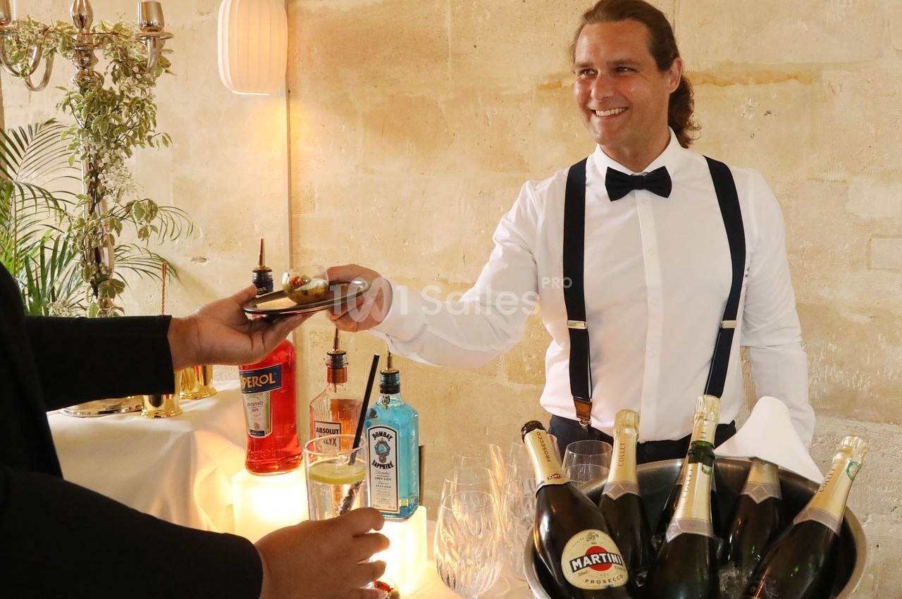 Un serveur souriant en chemise blanche et nœud papillon sert un amuse-bouche lors d'une réception avec boissons.