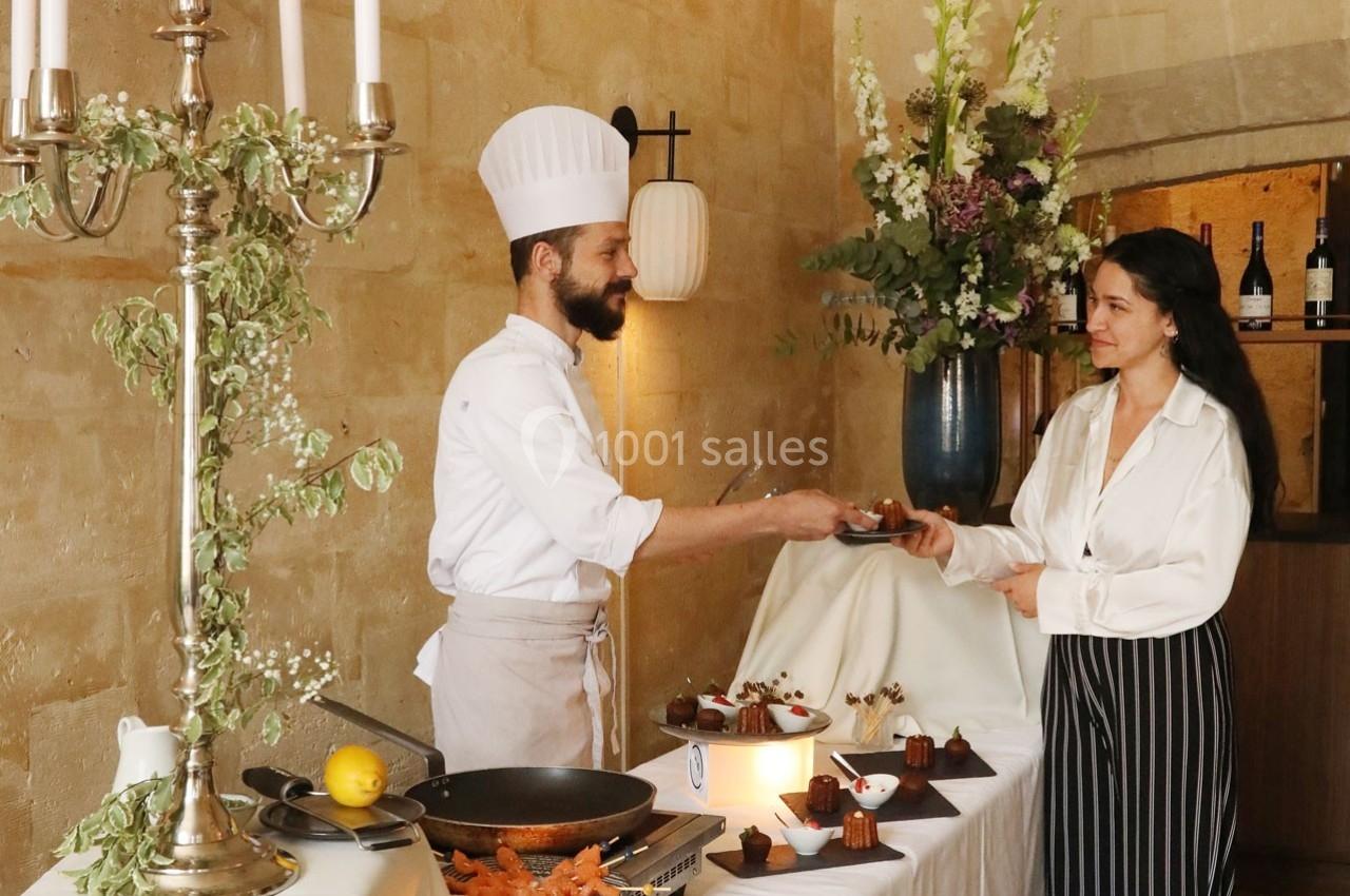Un chef sert un dessert à une femme dans un cadre élégant avec bougies, fleurs et présentations culinaires.