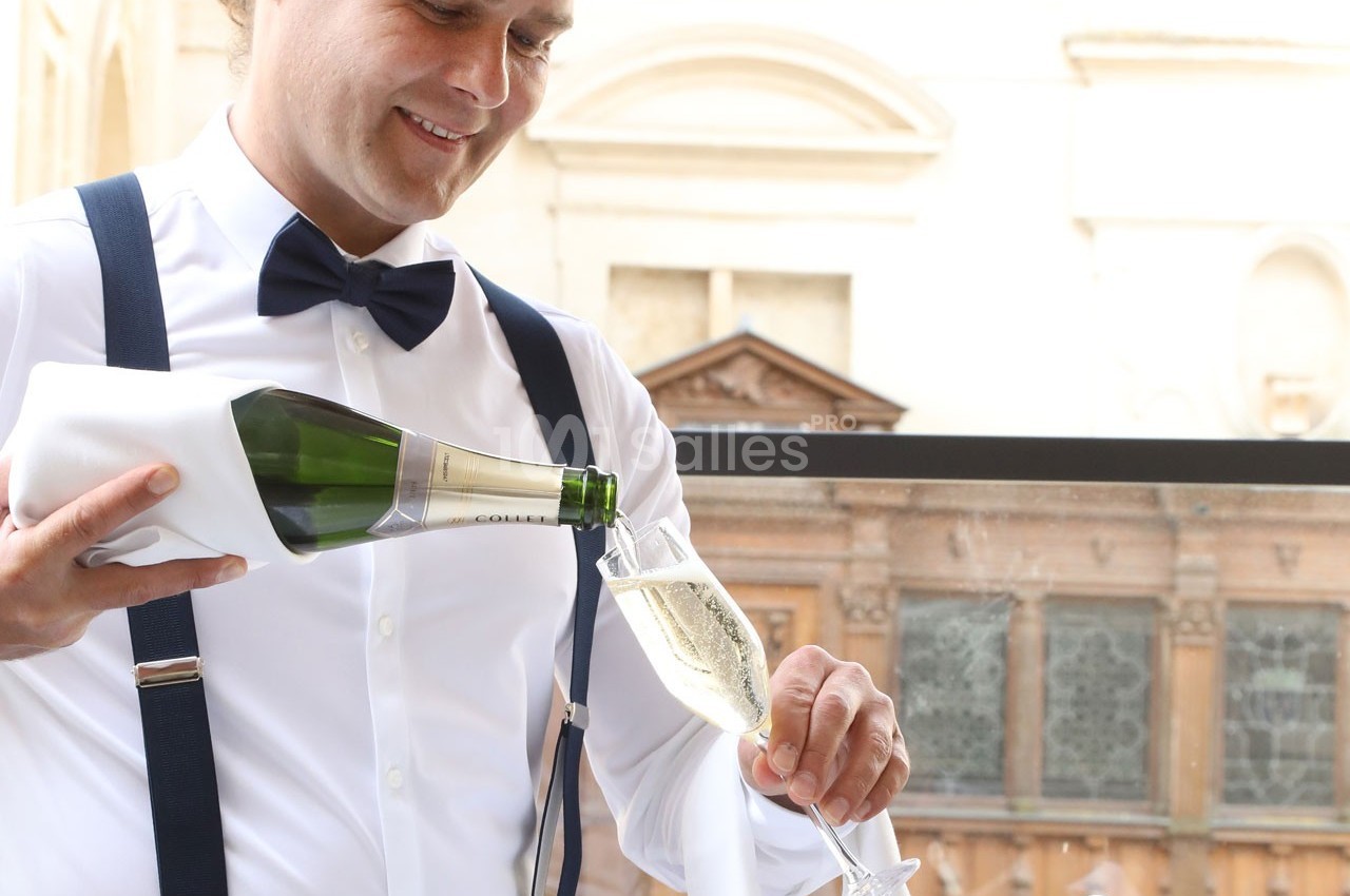 Un serveur en chemise blanche et nœud papillon verse du champagne dans une flûte sur une terrasse élégante.