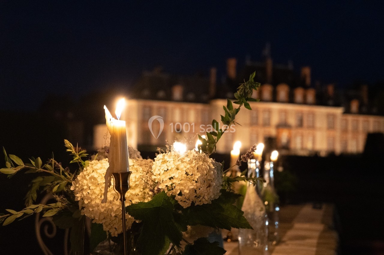 Bougies allumées et bouquets de fleurs blanches décorent une table en extérieur, avec un château éclairé en arrière-plan.