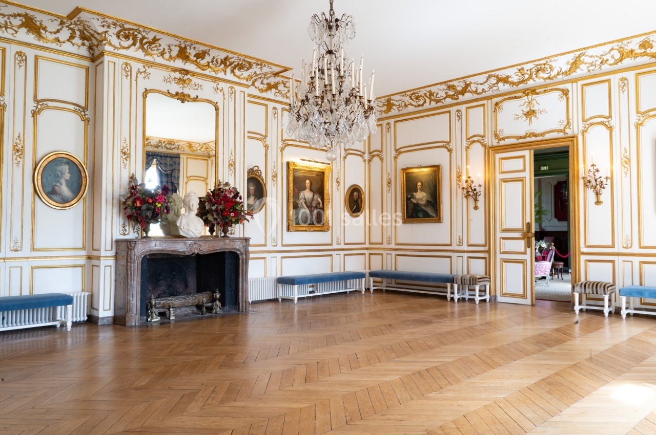 Salle élégante avec parquet en bois, moulures dorées, cheminée ornée, lustre central et portraits accrochés aux murs.