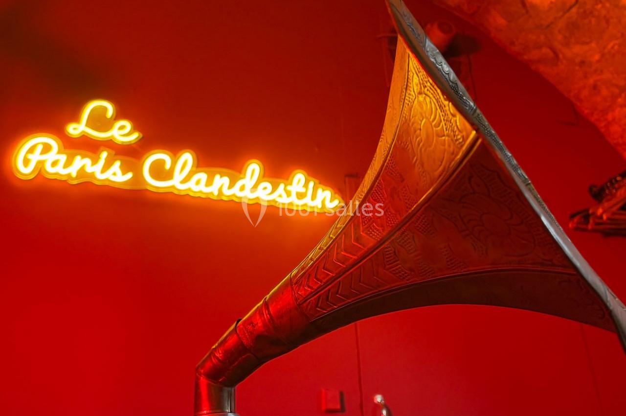 Phonographe ancien en métal gravé devant un néon orange indiquant ’Le Paris Clandestin’ sur un mur rouge.