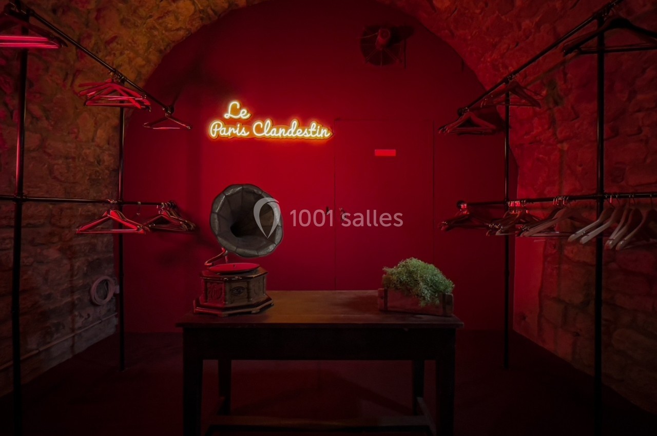 Salle en pierre éclairée de rouge avec un gramophone sur une table, des portants vides et une enseigne lumineuse.