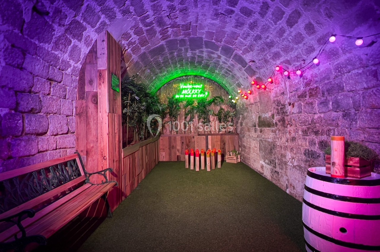 Allée voûtée en pierre éclairée par des lumières colorées, avec banc, tonneau et jeu de quilles sur gazon artificiel.