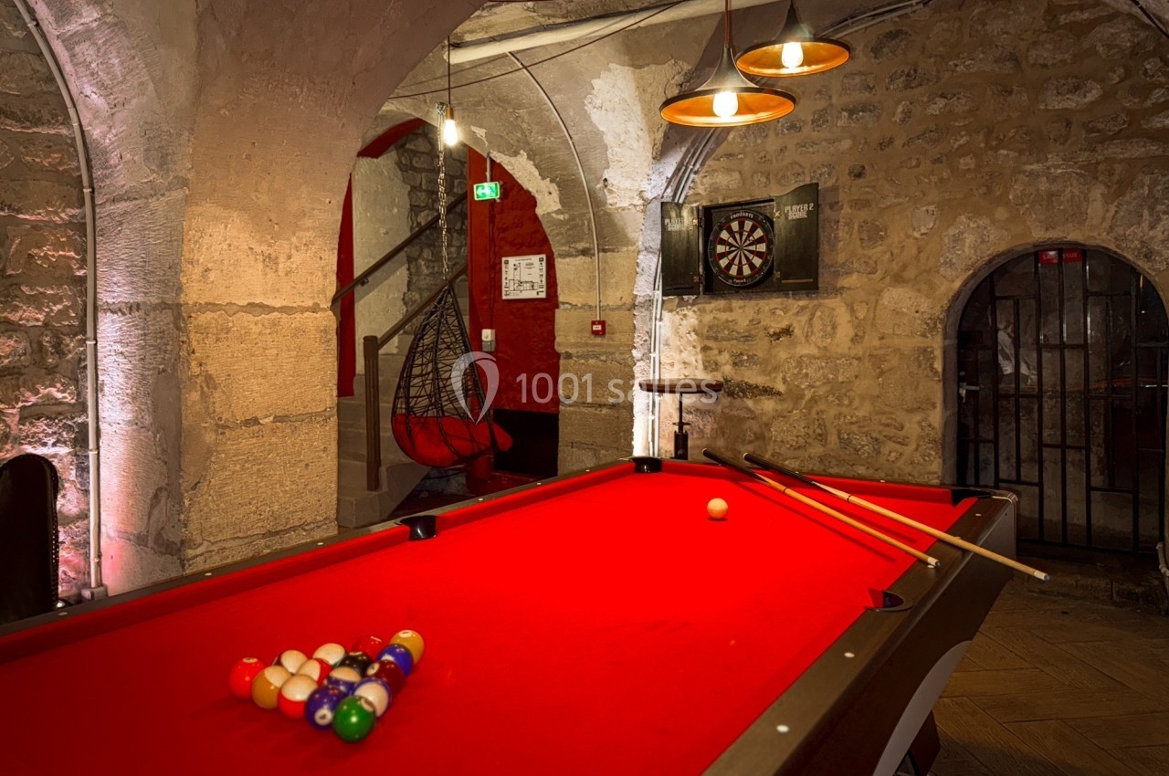 Table de billard rouge avec des queues et des boules colorées, dans une pièce en pierre avec un jeu de fléchettes au mur.