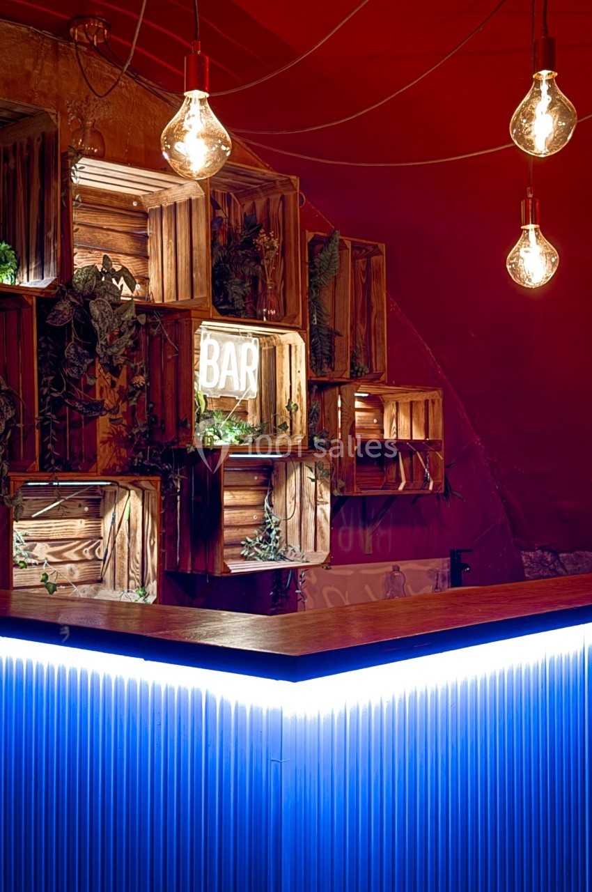 Comptoir de bar éclairé par des néons bleus, entouré de caisses en bois décorées de plantes et d'ampoules suspendues.