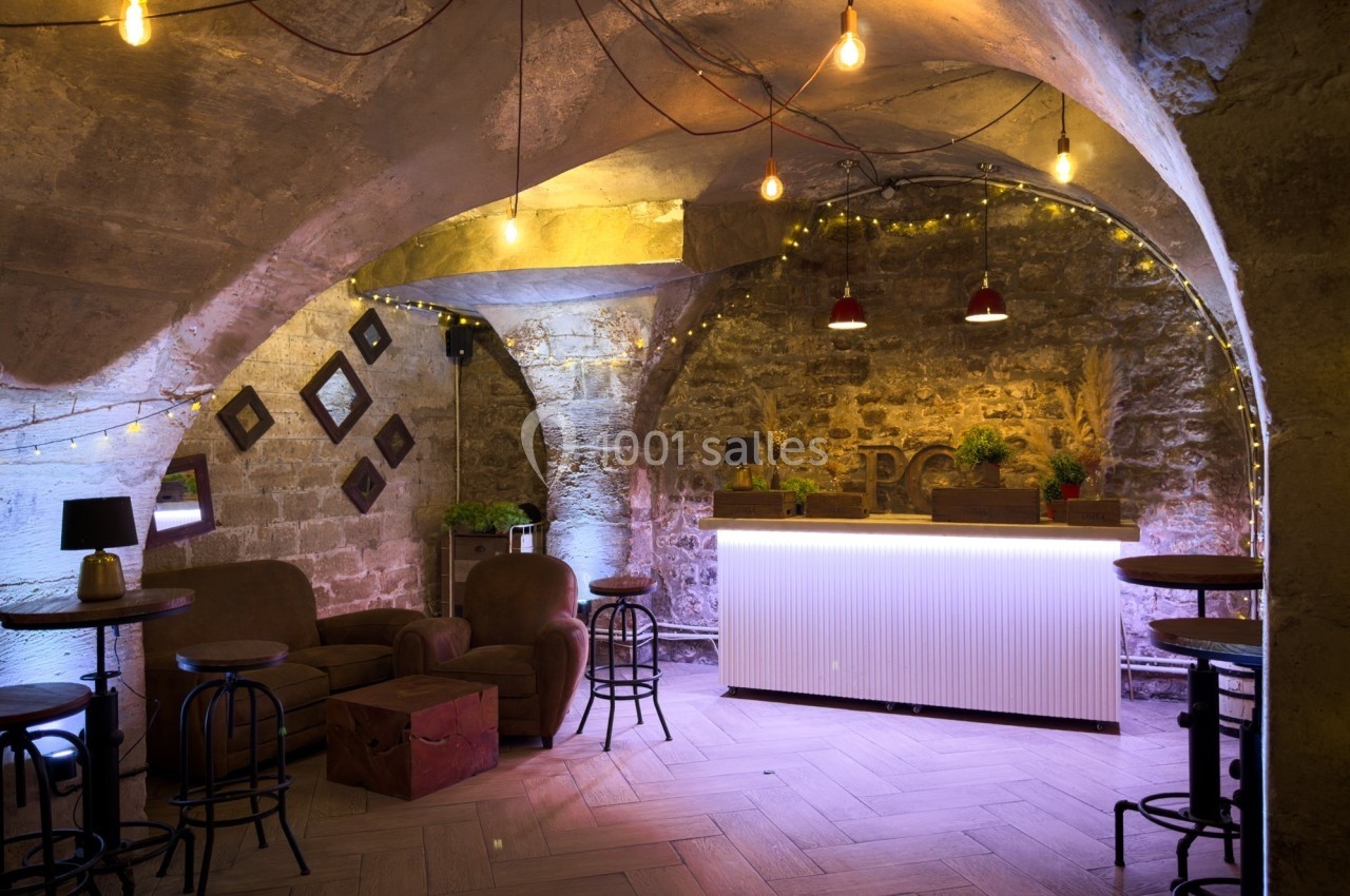 Salle voûtée en pierre avec un bar éclairé, des fauteuils, des tabourets et des guirlandes lumineuses suspendues.