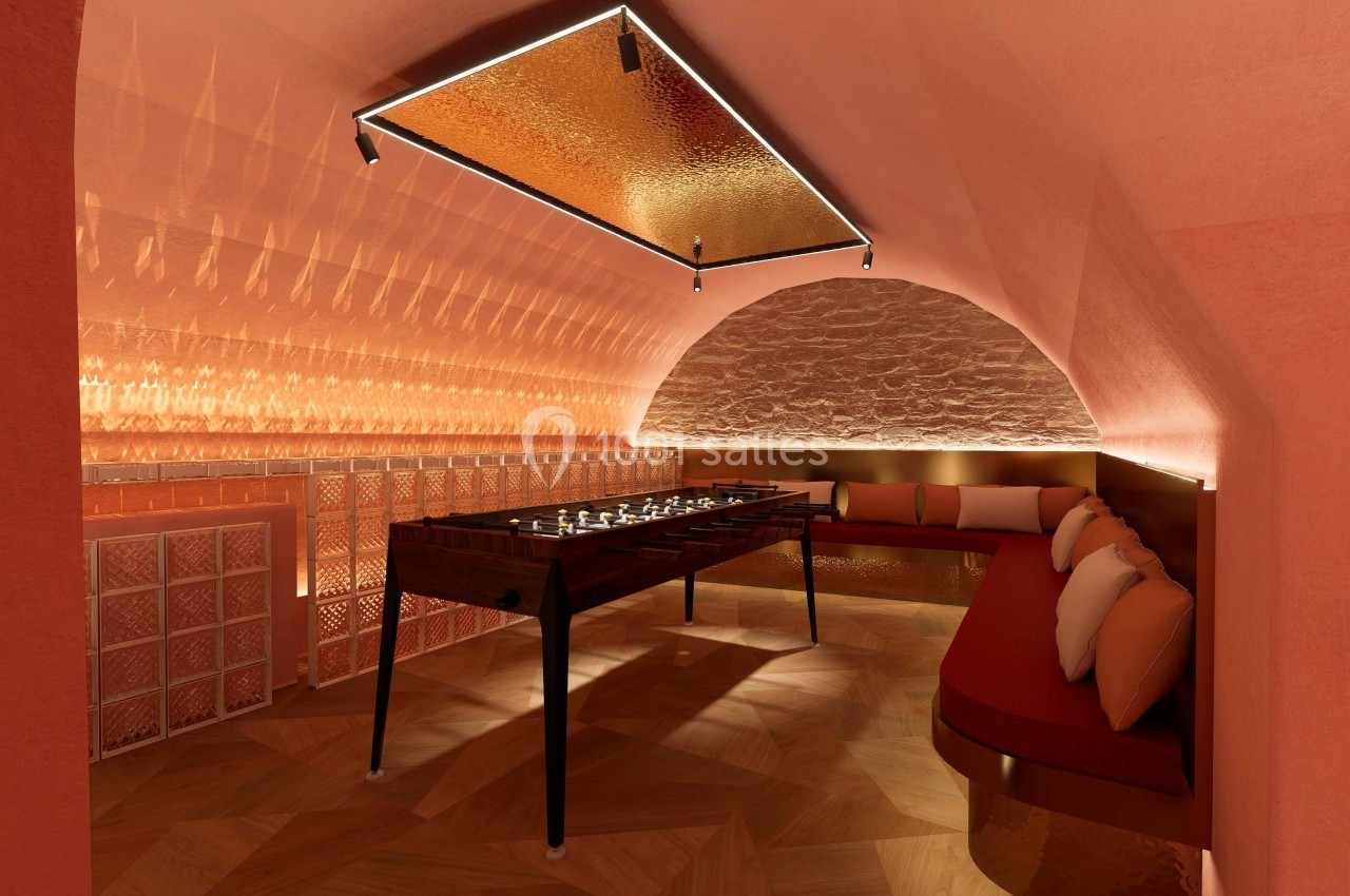 Salle voûtée avec éclairage chaleureux, baby-foot en bois au centre et banquette arrondie avec coussins colorés.