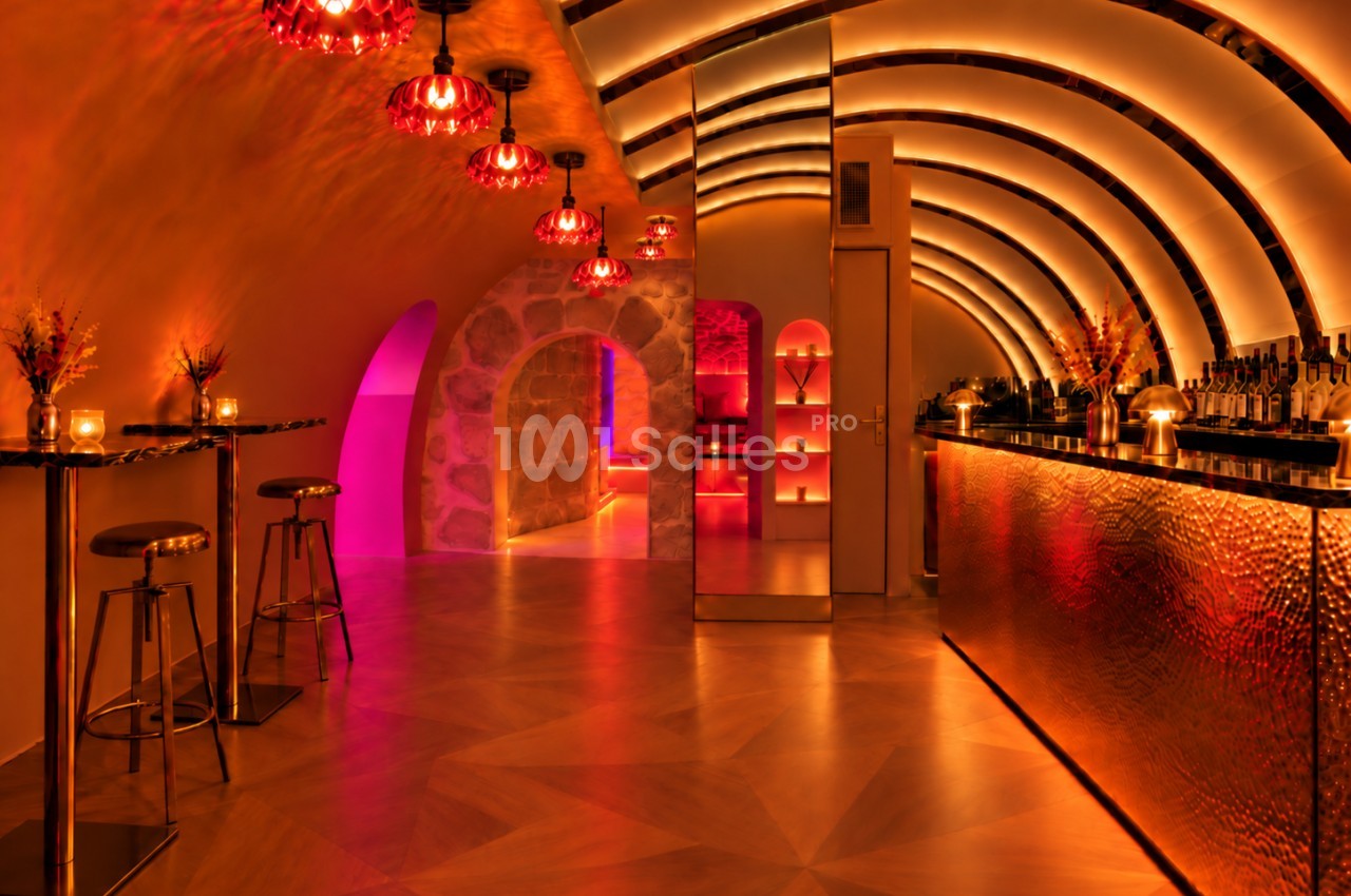 Bar voûté aux lumières orangées et roses, avec suspensions rouges, tabourets hauts, comptoir en cuivre martelé et arches éclairées.