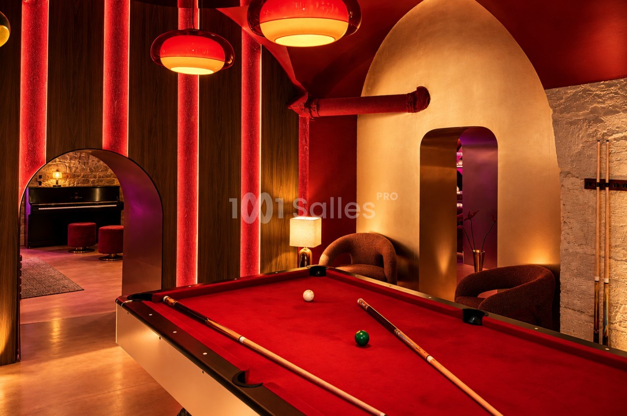 Table de billard au tapis rouge dans une salle aux murs en bois et lumières rouges, avec des fauteuils bouclés et une arche menant à un piano.