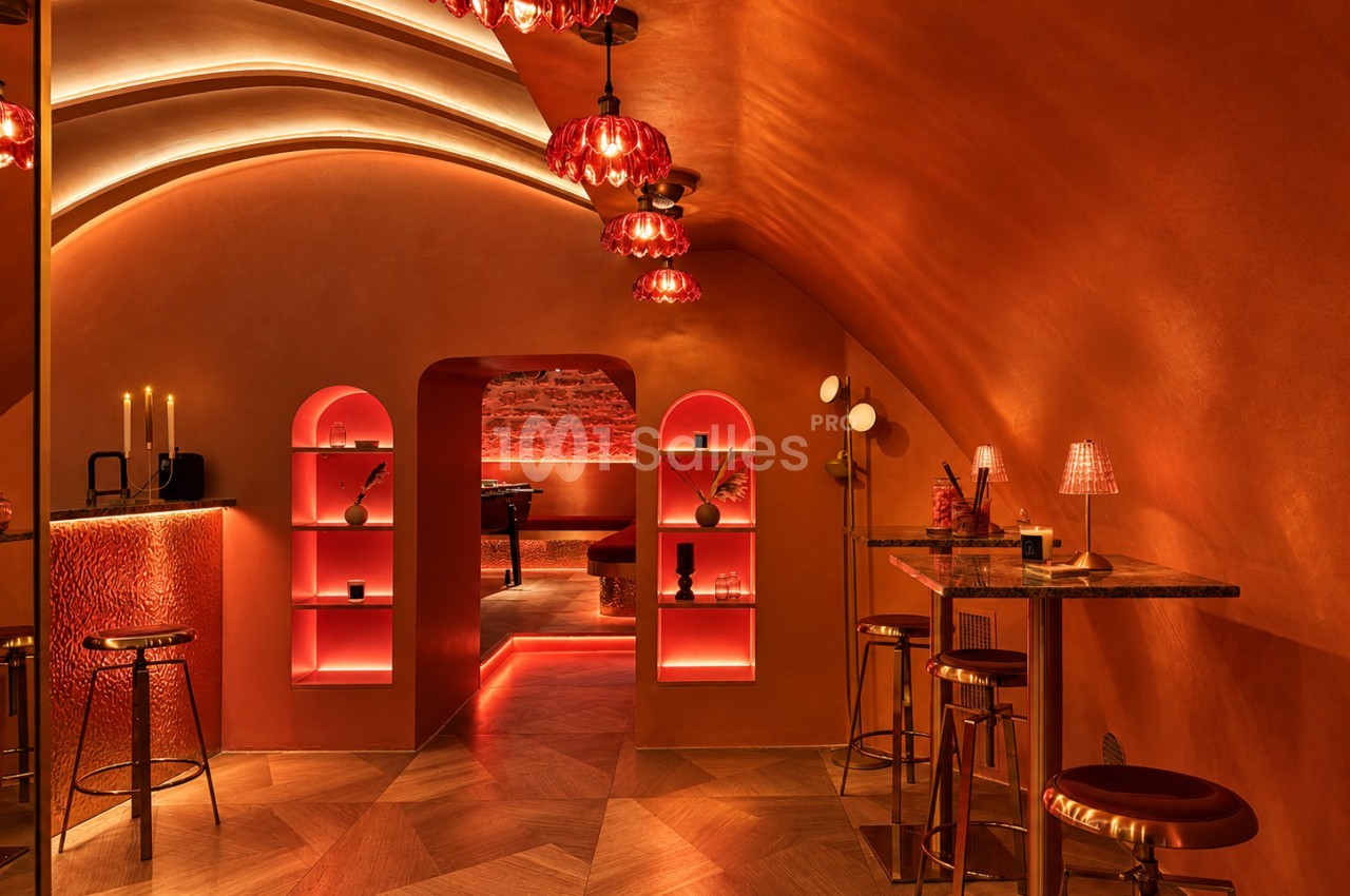 Salle aux murs ocre et lumière rouge-orangée, avec niches en arche éclairées, lustres en verre rouge, mange-debout en marbre et tabourets dorés.