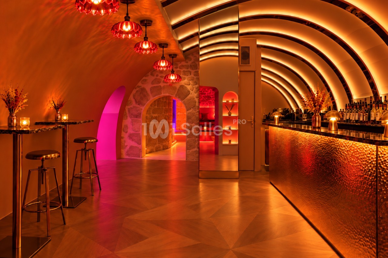 Bar en sous-sol aux voûtes arrondies éclairées en orangé, avec suspensions rouges, hauts tabourets et comptoir métallique martelé.