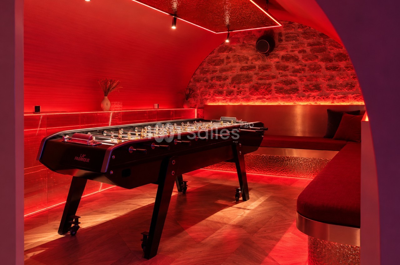 Salle voûtée à éclairage rouge avec un baby-foot noir au centre, banquettes en bordure et mur en pierre apparente.