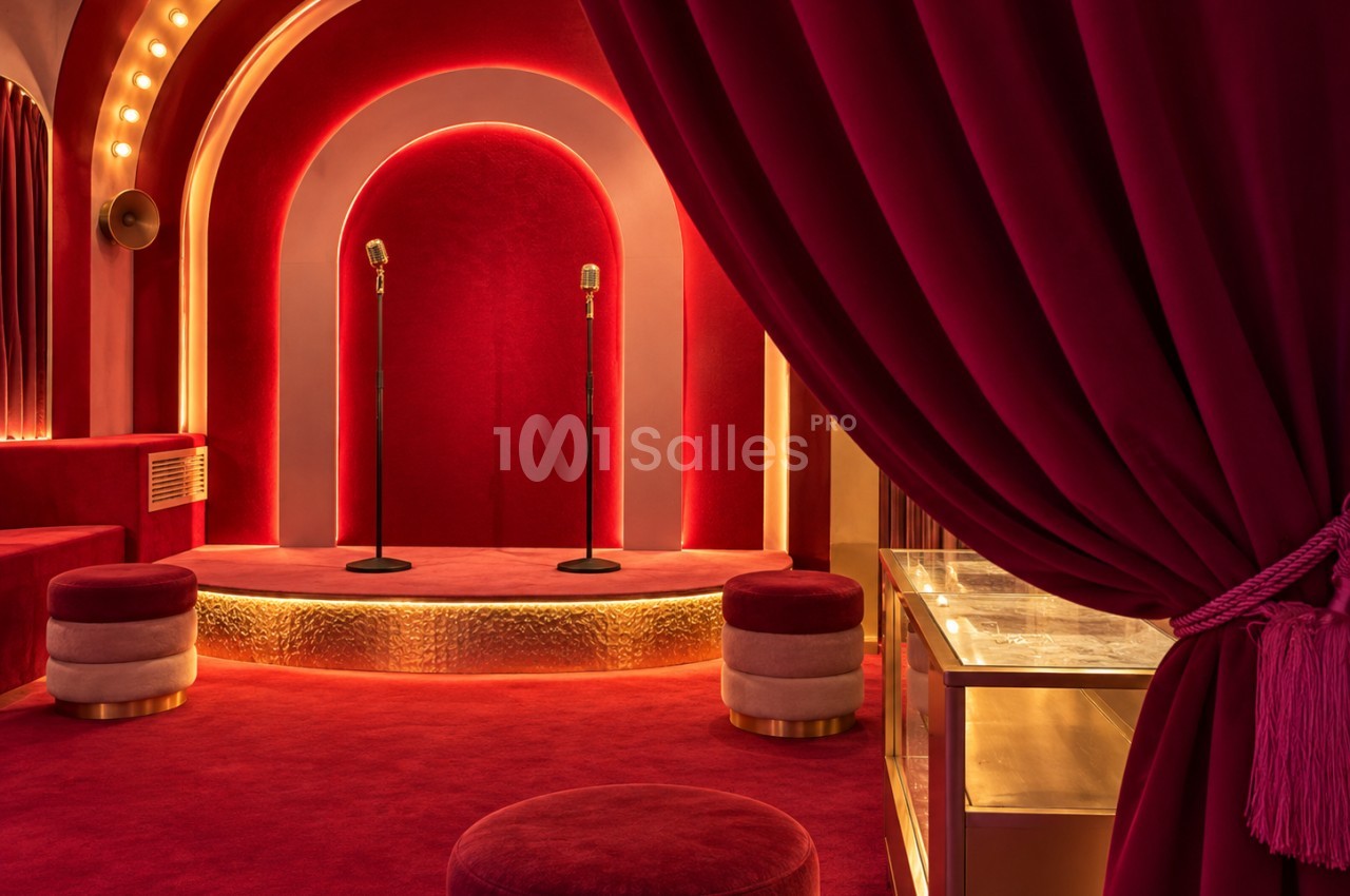 Scène rouge et dorée sous arches lumineuses, avec deux micros vintage sur un petit podium, des poufs capitonnés et un rideau de velours au premier plan.