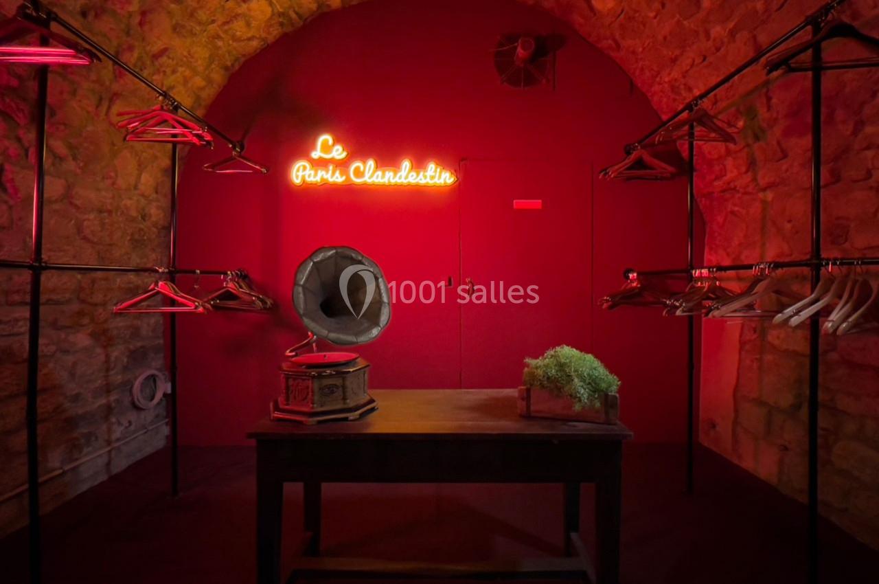 Salle en pierre éclairée de rouge avec un gramophone, une table, des portants vides et un néon ’Le Paris Clandestin’.