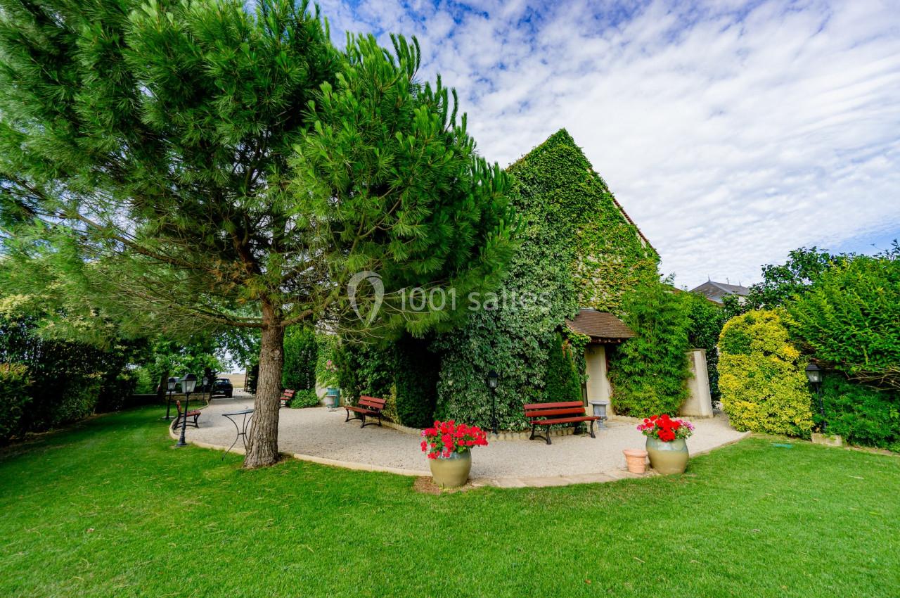 Maison recouverte de lierre entourée d'un jardin verdoyant avec bancs, pots de fleurs et arbres sous un ciel partiellement…