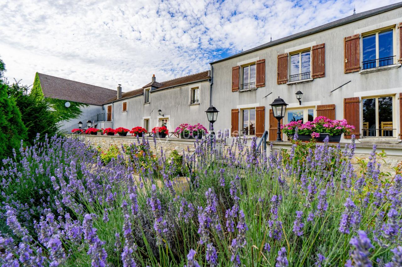 Façade d'une maison avec volets en bois, jardinières fleuries et lavandes au premier plan sous un ciel partiellement nuageux.